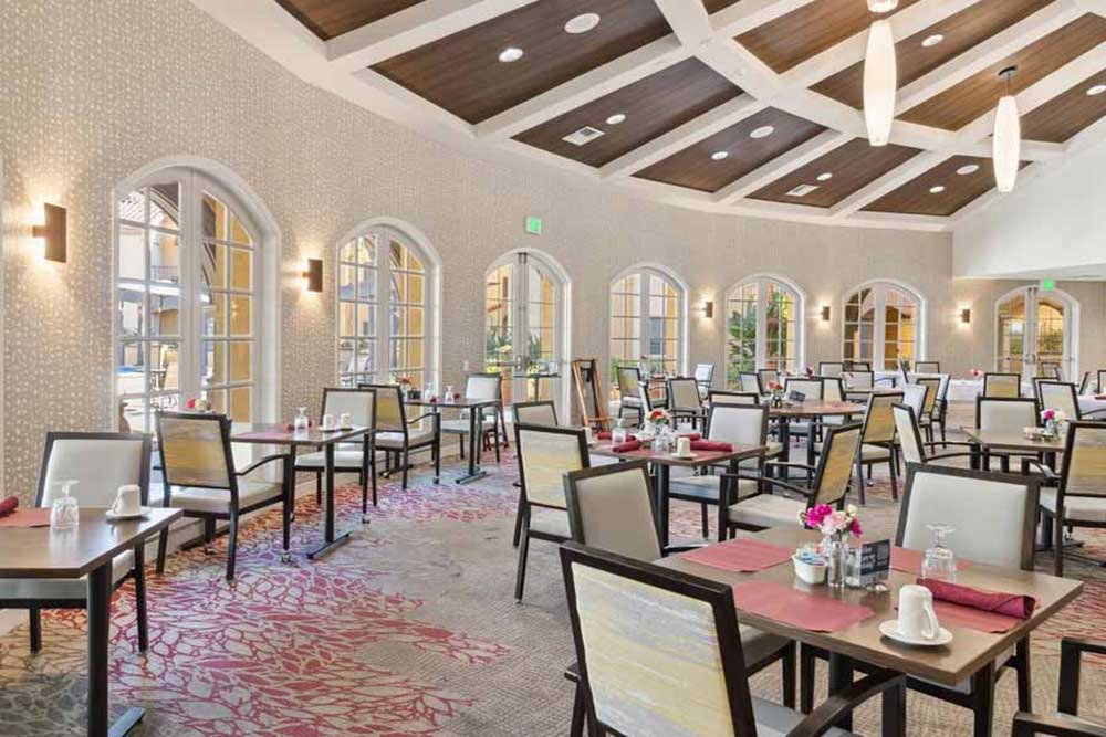 A spacious, elegant dining room with large arched windows, neutral walls, wooden ceiling beams, and neatly set tables with beige chairs, red placemats, and flower centerpieces. Natural light fills the room.