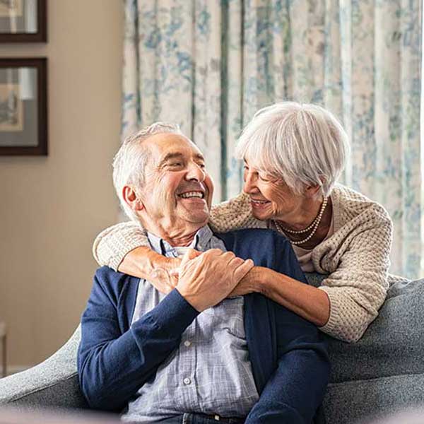 An elderly couple sits together on a couch, smiling warmly. The woman hugs the man from behind, and they look at each other fondly. The background shows patterned curtains and framed pictures on the wall.