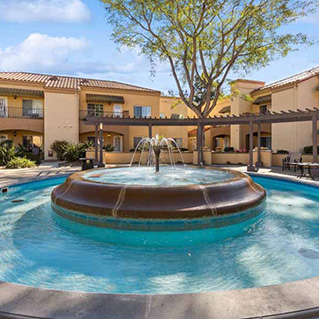 A large round fountain with water jets is in the center of a courtyard, surrounded by a paved area, benches, and a tree. In the background are two-story, beige buildings with balconies and tiled roofs.