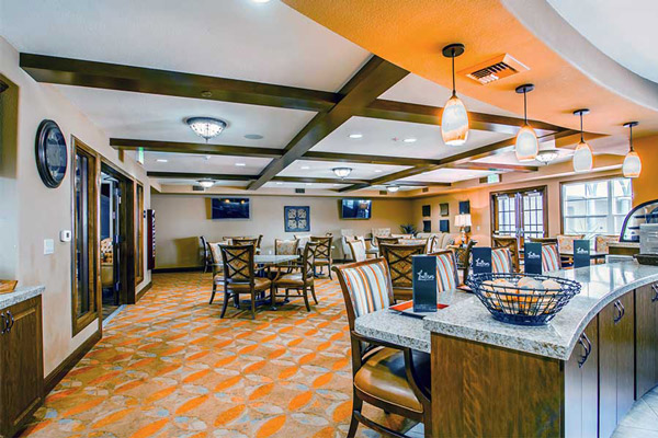 A spacious, brightly lit dining area with wooden tables and chairs, patterned orange carpet, pendant lights, and a counter with menus and a bread basket. Large windows let in natural light.