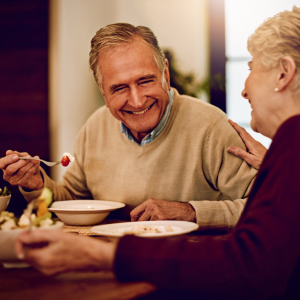 An older man and woman sit at a dining table, smiling and enjoying a meal together. The man holds a spoon with food, and the woman rests her hand on his arm in a warm, friendly gesture.