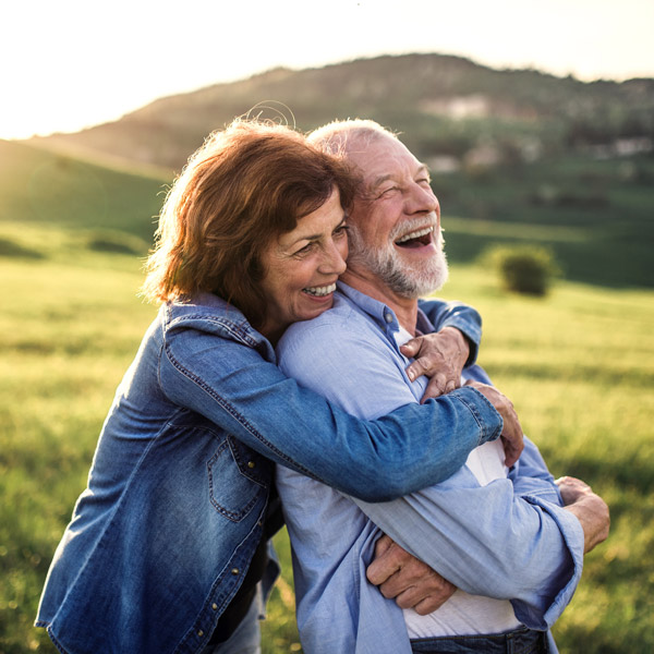 An older couple stands outdoors in a green field, smiling and laughing as the woman hugs the man from behind. Both wear casual clothes and look happy under the warm sunlight with hills in the background.