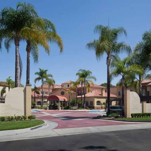 A gated entrance to a Mediterranean-style building with red tile roofs, surrounded by tall palm trees, landscaped greenery, and a red-brick driveway under a clear blue sky.