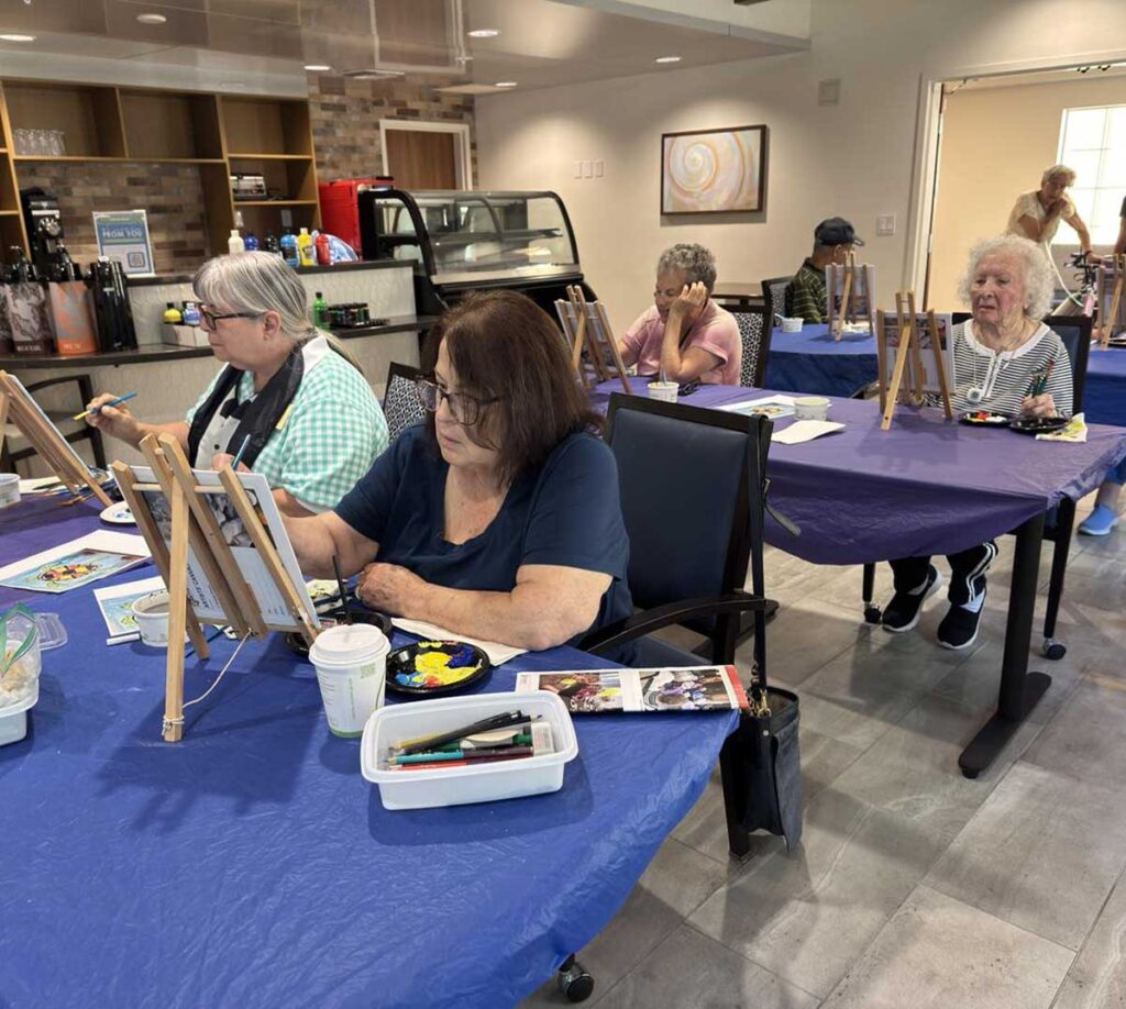 A group of older adults sit at tables covered with blue cloths, painting on canvases in a bright, modern room. Art supplies and coffee cups are on the tables, and the atmosphere appears focused and creative.