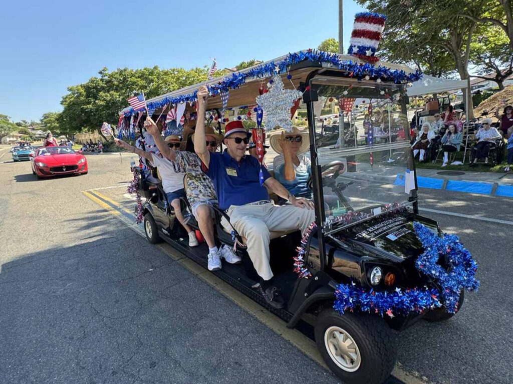 A decorated golf cart with people waving American flags and wearing patriotic attire drives down a street during a parade. Red, white, and blue decorations cover the cart, and spectators watch from the sidewalk.