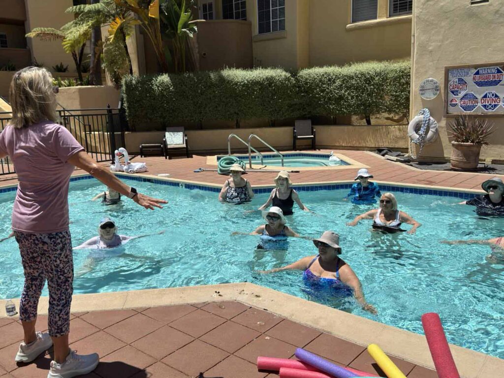 A group of older adults wearing hats participates in a water aerobics class in an outdoor pool, led by an instructor standing poolside. Colorful pool noodles are on the ground nearby.