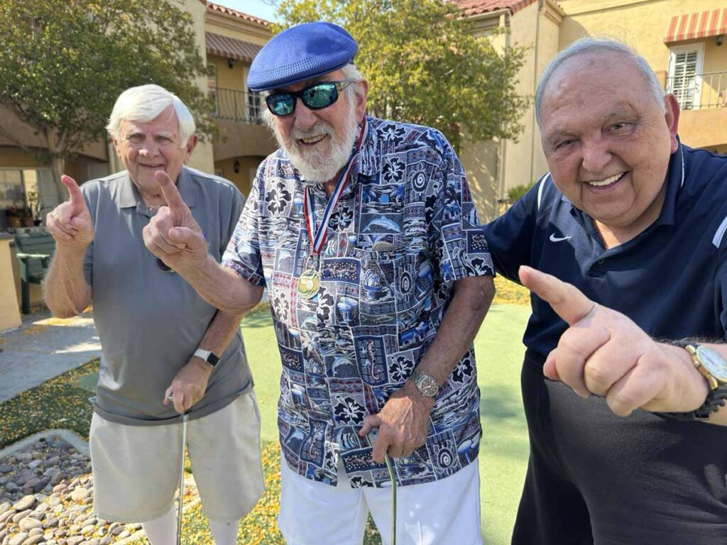 Three older men smiling and holding up one finger each, standing outdoors on a sunny day. The man in the middle wears a blue hat, sunglasses, a patterned shirt, and a medal. The others wear casual shirts and shorts.