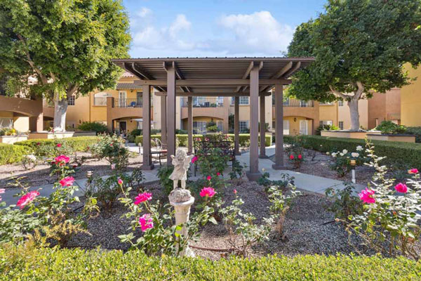 A landscaped garden with blooming pink and white roses, a small statue, and a covered pergola. Yellow buildings and tall green trees surround the courtyard under a partly cloudy sky.