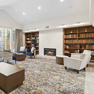 A spacious, modern living room with a patterned rug, armchairs, wooden tables, built-in bookshelves filled with books, a fireplace, and large windows letting in natural light.