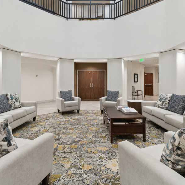 A spacious, modern lobby with several light-colored sofas arranged around a coffee table on a patterned rug. The area is well-lit with white walls and double wooden doors in the background.