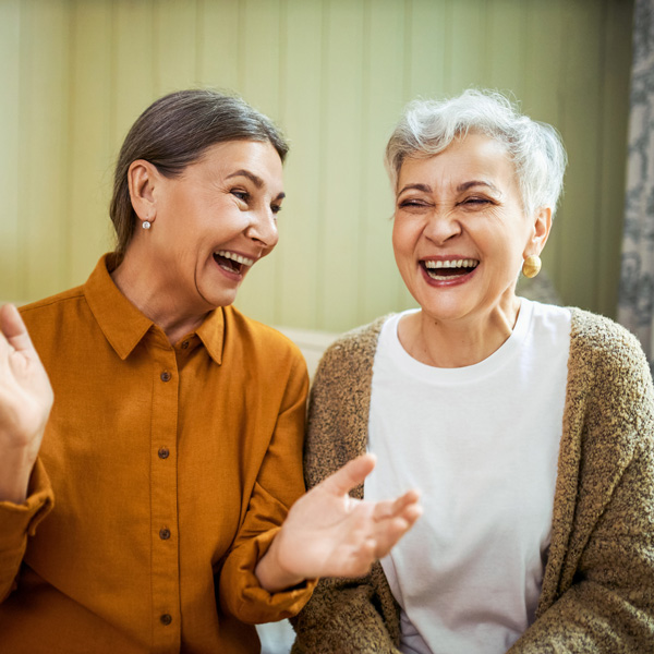 Two older women sitting close together indoors, laughing and smiling warmly. One wears a mustard-colored shirt, the other a white top with a beige cardigan. They appear to be enjoying a happy conversation.