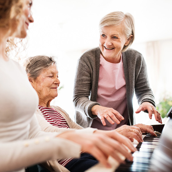 Three older women smiling and playing the piano together indoors, with one woman standing and two women sitting, sharing a joyful moment.