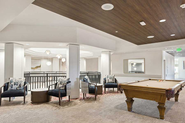 A modern lounge area with a pool table, four upholstered chairs with patterned pillows, a round table, and neutral decor. The space features a wood-paneled ceiling section and a large mirror on the wall.