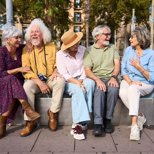 Five older adults sit on a bench outdoors, smiling and laughing together. They appear relaxed and happy, dressed in casual clothing, enjoying a sunny day with palm trees in the background.