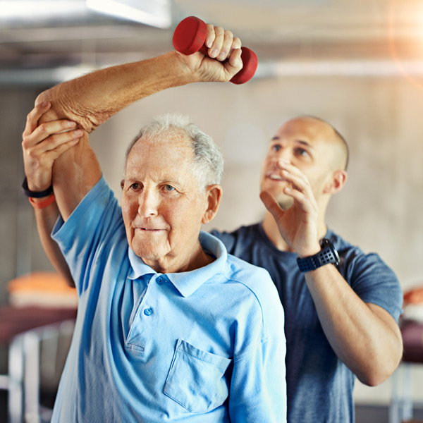 An older man in a blue shirt lifts a red dumbbell overhead with assistance from a younger man, likely a physical therapist, in a gym or rehabilitation setting.