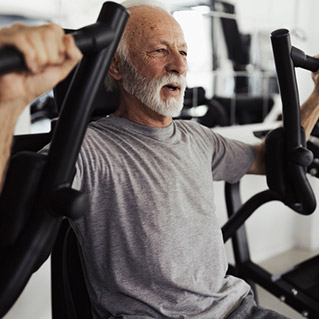 An older man with a gray beard and short hair works out on a chest press machine at a gym. He is wearing a gray t-shirt and looks focused while exercising.