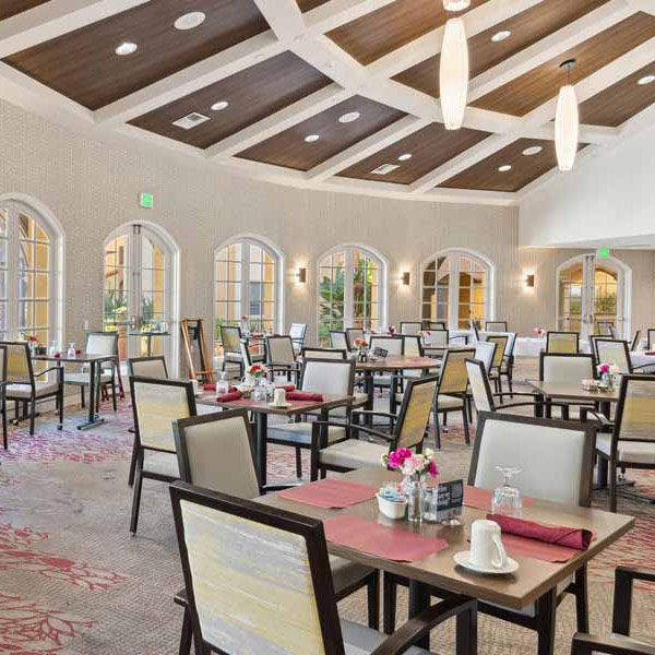 A spacious, elegant dining room with neatly arranged tables and chairs, set with plates, glasses, and napkins. Large arched windows line the wall, letting in natural light. The ceiling features wooden beams and modern light fixtures.