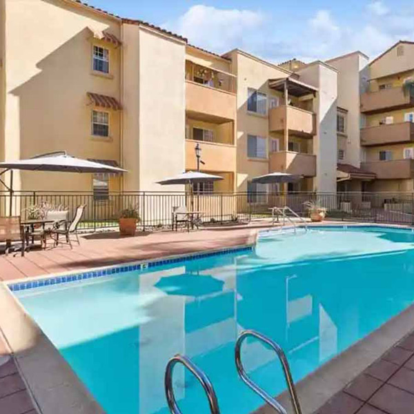 A clear outdoor swimming pool with metal handrails, surrounded by lounge chairs, tables with umbrellas, and potted plants, in front of a multi-story beige apartment building.