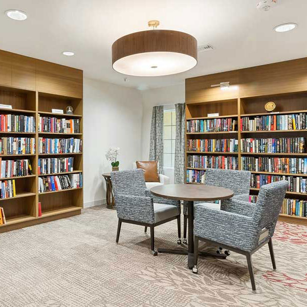 A cozy library room with three patterned chairs around a round table, surrounded by bookshelves filled with books. The room has neutral tones, a large ceiling light, and a window with sheer curtains.