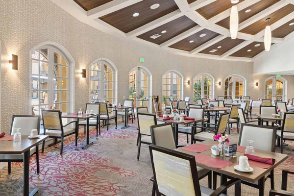 A bright, spacious dining room with large arched windows, neatly arranged tables with red placemats, cushioned chairs, modern lighting fixtures, and floral centerpieces on each table.