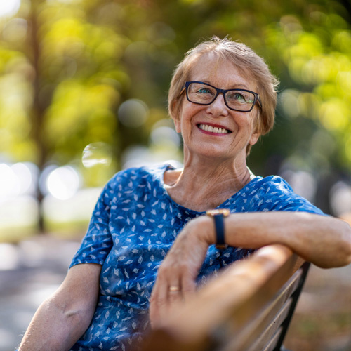 An older woman with short hair and glasses smiles while sitting on a park bench. She is wearing a blue patterned shirt and a watch, with sunlight and greenery in the blurred background.