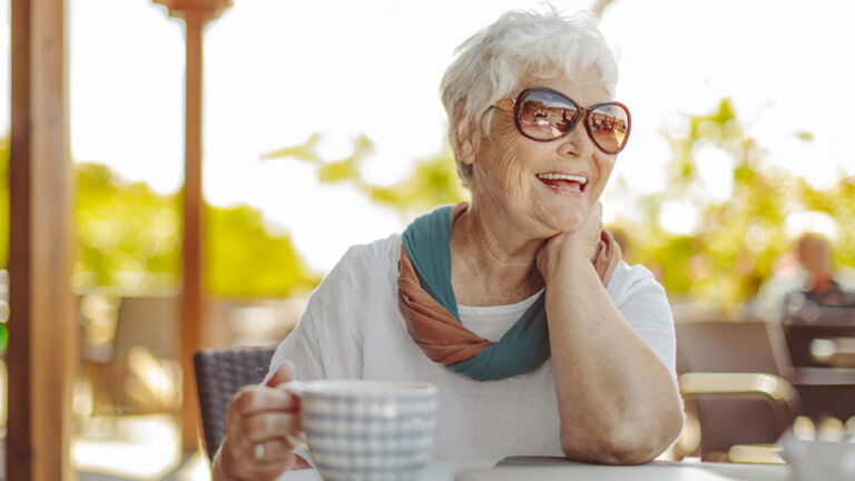 An elderly woman with short white hair and sunglasses smiles while sitting at an outdoor table, enjoying life in senior living. She holds a cup, wears a scarf and white shirt, with sunny blurred greenery and chairs in the background.