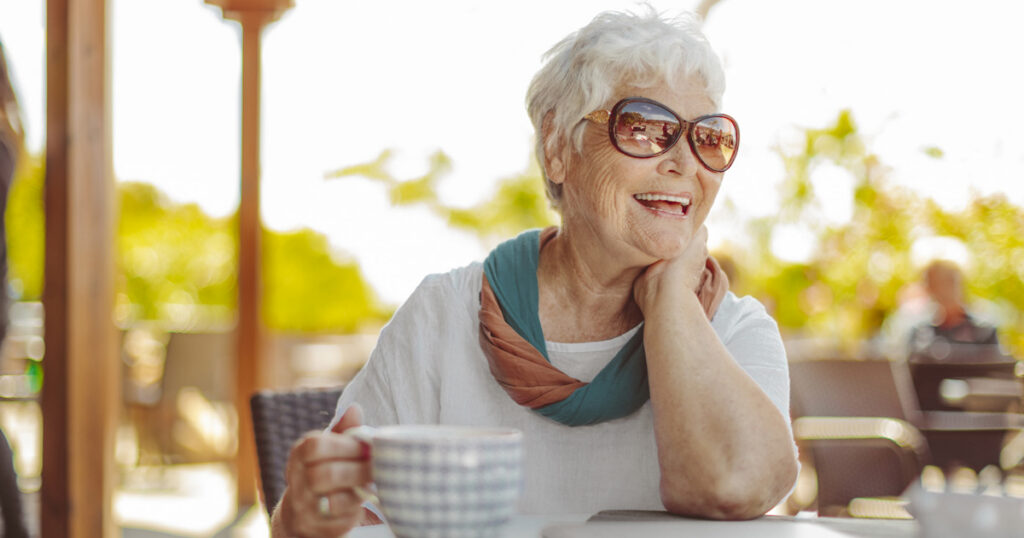 Elderly woman with white hair wearing large sunglasses and a scarf sits at an outdoor café table, smiling and holding a coffee cup, with sunlight and greenery in the background.