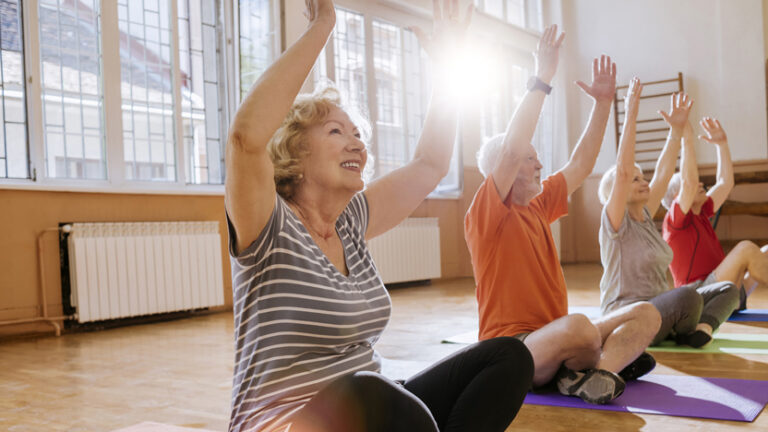 Four older adults sit on yoga mats in a sunlit room, raising their arms and smiling during a group exercise or yoga class. Large windows let sunlight stream in, creating a bright and cheerful atmosphere.