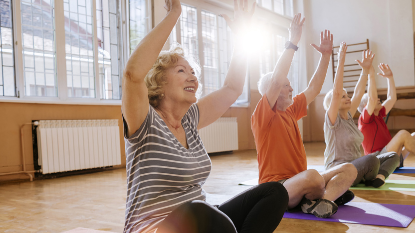 Four older adults sit on yoga mats in a sunlit room, raising their arms and smiling during a group exercise or yoga class. Large windows let sunlight stream in, creating a bright and cheerful atmosphere.